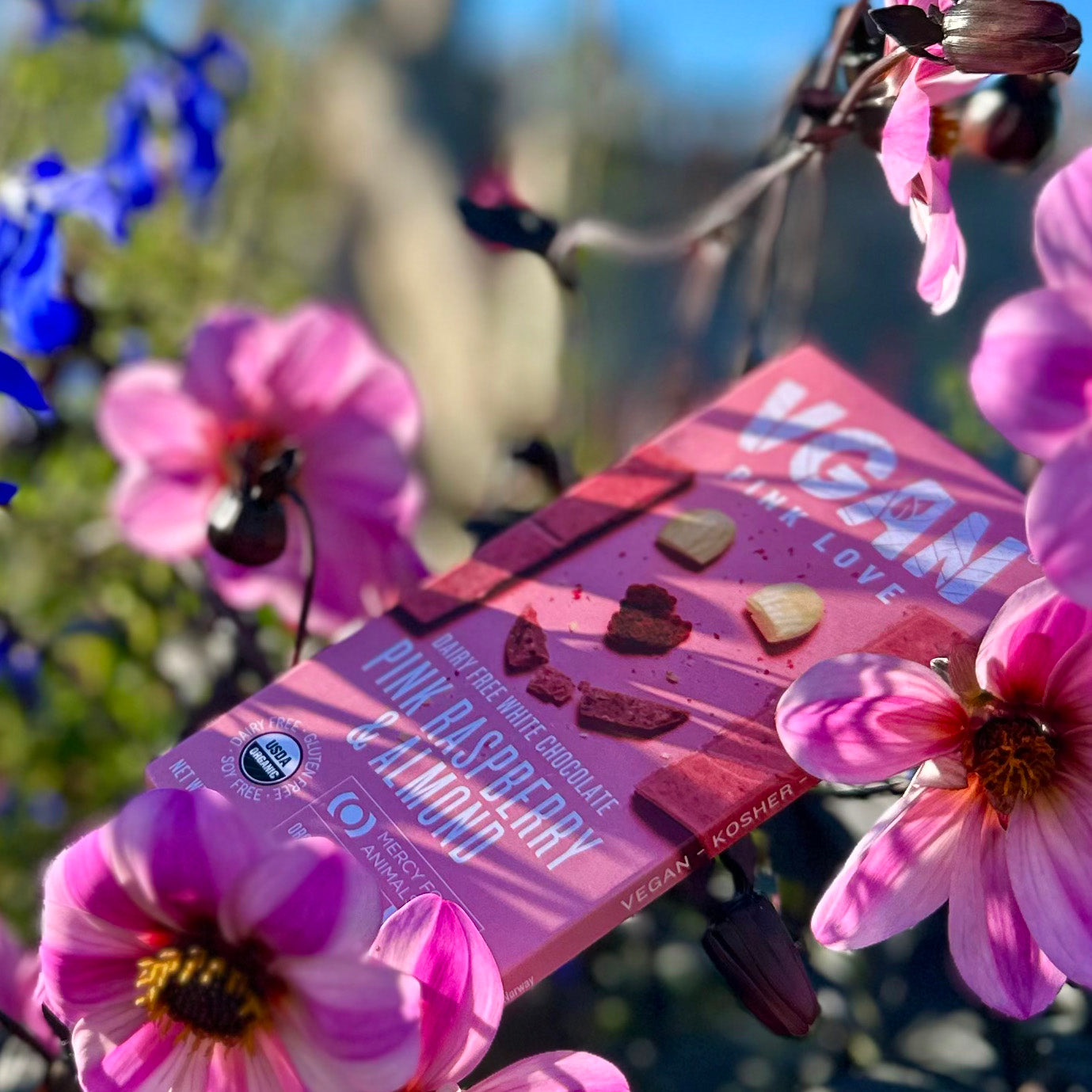 Vegan White Chocolate with Pink Raspberry & Almond laying between some beautiful Pink and Blue flowers